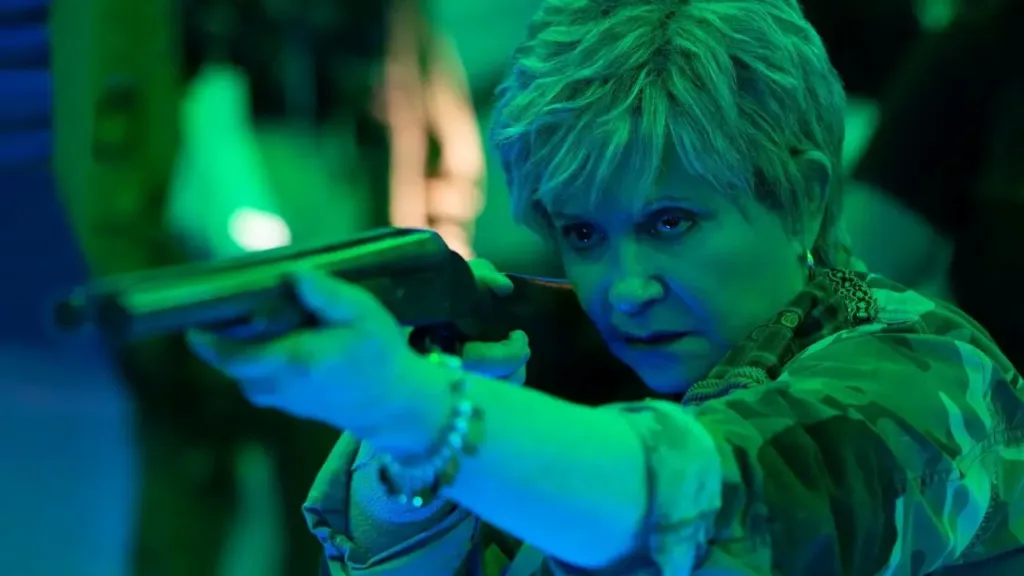 Μια κοντινή φωτογραφία της Adriana Barraza από την ταινία "Bingo Hell" του 2021, όπου κρατάει ένα τουφέκι και φοράει στρατιωτικό τζάκετ, με πράσινο φωτισμό.