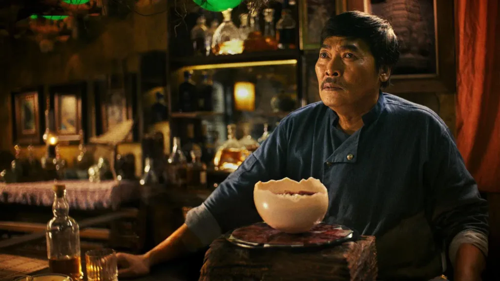 Στιγμιότυπο από τη σειρά τρόμου 'Devil's Diner' (2025): Μυστηριώδης σεφ σε σκοτεινό εστιατόριο με περίτεχνο πιάτο σε σπασμένο αυγό πάνω σε δίσκο, ανάμεσα σε μπουκάλια και ποτήρια.