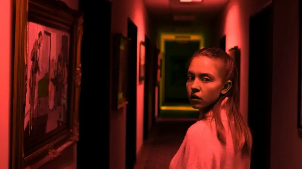 Νέο κορίτσι με μακριά κόκκινα μαλλιά κοιτάζει επίμονα κάτω από κόκκινο φως σε σκοτεινό διάδρομο με πλαίσιο στο φόντο, σκηνή από το Nocturne 2020.