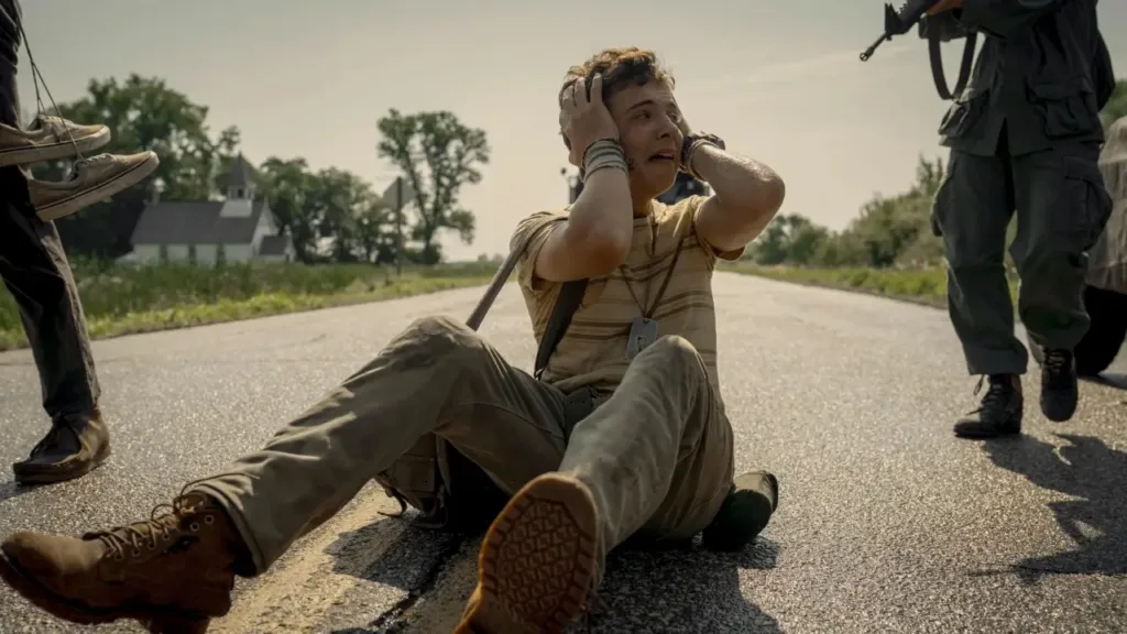 Άνδρας κάθεται τρομαγμένος στη μέση του δρόμου, ενώ στρατιώτες τον περικυκλώνουν. Στιγμιότυπο από την ταινία "The Long Walk" (2025).﻿