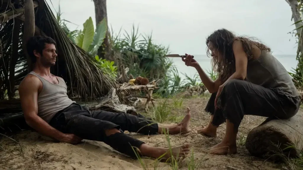 Γυναίκα ναυαγός κρατά μαχαίρι και το δείχνει σε τραυματισμένο άντρα που κάθεται στο έδαφος δίπλα σε πρόχειρο καταφύγιο από φύλλα στην άκρη της παραλίας, μέσα σε πυκνή βλάστηση.