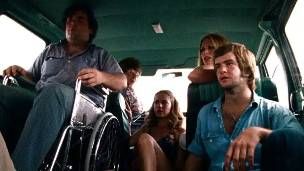 Ομάδα πέντε νεαρών φίλων στριμωγμένοι μέσα σε βαν, με έναν άντρα σε αναπηρικό αμαξίδιο στο μπροστινό μέρος, σε χαλαρή στιγμή στην αρχή του ταξιδιού τους πριν αρχίσει ο εφιάλτης στο The Texas Chain Saw Massacre (1974).