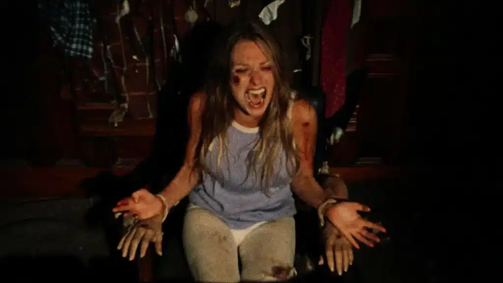 Τρομαγμένη νεαρή γυναίκα κάθεται δεμένη σε καρέκλα, χτυπημένη και αιματωμένη, περιτριγυρισμένη από κρεμασμένα ρούχα και μακάβρια διακοσμητικά χέρια, καθώς παγιδεύεται στο σπίτι των δολοφόνων στο The Texas Chain Saw Massacre (1974).