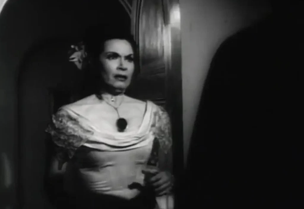 Γυναίκα σε ασπρόμαυρο πλάνο στέκεται σε σκοτεινό διάδρομο, φορώντας βραδινή φόρμα εποχής με κολιέ και κρατώντας μαχαίρι στο χέρι, έτοιμη για επίθεση. Σκηνή από την ταινία «Curse of the Stone Hand».