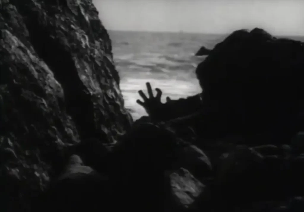 Σιλουέτα σώματος κείτεται ανάμεσα σε βράχια δίπλα στη θάλασσα, με ένα χέρι τεντωμένο προς τα πάνω σε ασπρόμαυρο, ομιχλώδες πλάνο τρόμου.