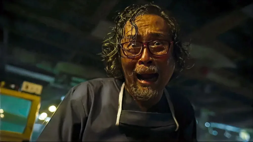 Άντρας με ποδιά εργαστηρίου και ατημέλητα μαλλιά σκύβει απειλητικά προς την κάμερα σε μισοσκότεινο, βιομηχανικό χώρο, σαν τρελός επιστήμονας από βιντεοκασέτα τρόμου.