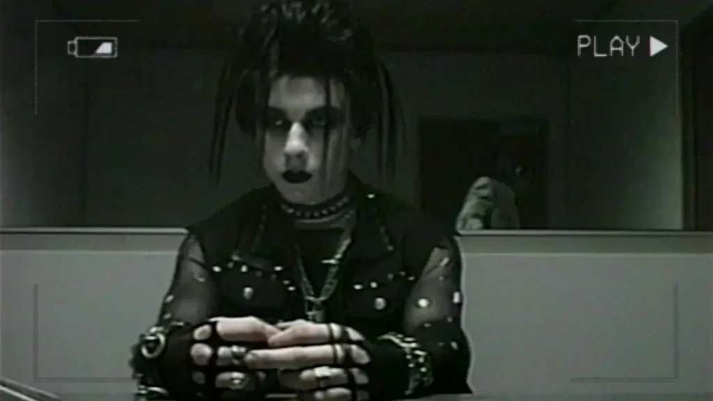Νεαρό άτομο με goth ρούχα και καρφιά κάθεται σε αίθουσα ανάκρισης, τραβηγμένο από βίντεο κασέτας, ενώ στο βάθος διακρίνεται τρομακτική φιγούρα στην πόρτα. Σκηνή από την ταινία «V/H/S/85».